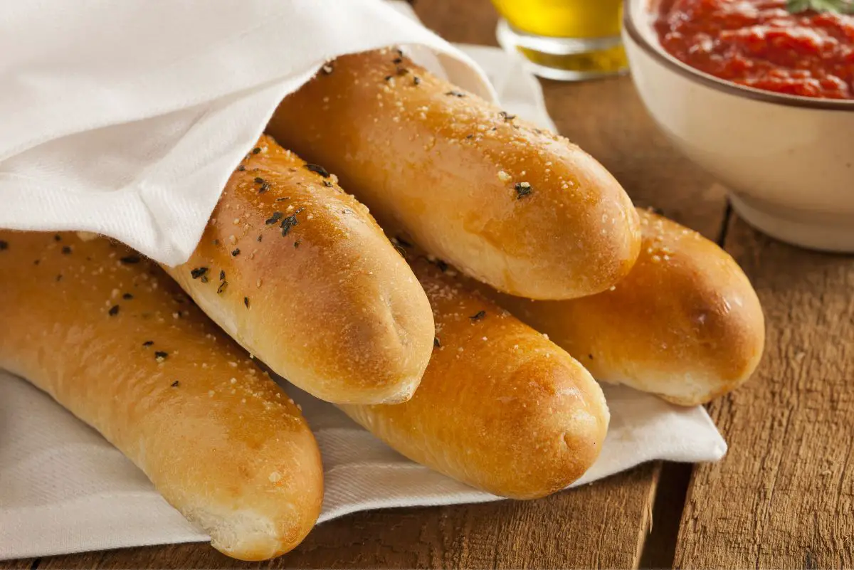 Three freshly baked, fluffy breadsticks with a glossy, golden-brown crust, speckled with herbs and salt, neatly lined up on a white linen cloth on a textured wooden table surface, accompanied by a creamy white bowl of marinara sauce in the soft-focused backdrop.