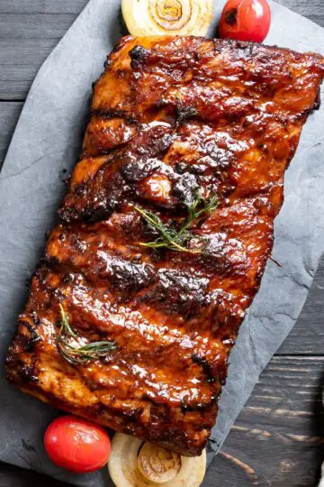 Close-up of BBQ ribs on slate with a rich sauce, onions, and tomatoes beside, with rosemary on top.