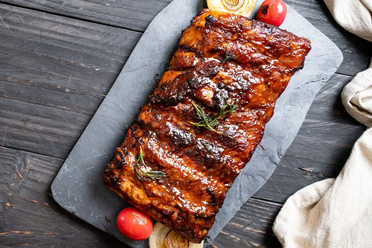 BBQ ribs on parchment over a slate, with charred onions, tomatoes, and rosemary, on a rustic wood background.