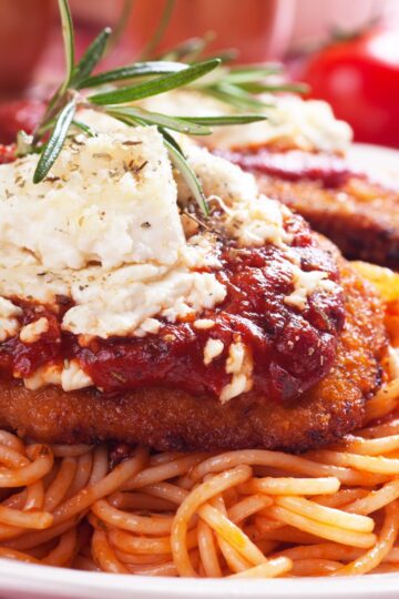 This is a close-up photograph highlighting the textures and colors of the Simple Italian Chicken Parmesan dish. A golden-brown breaded chicken breast, smothered in a rich red marinara sauce and topped with a generous portion of crumbled white Monterey Jack cheese, sits atop a twist of al dente spaghetti. The dish is garnished with a sprig of fresh rosemary, adding a pop of green against the warm tones of the chicken and pasta.