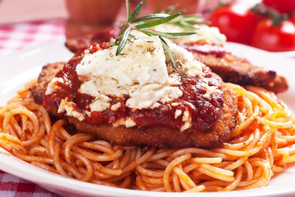The image presents a well-composed photo of Simple Italian Chicken Parmesan served on a white plate. The plate features a breaded chicken breast covered in a vibrant tomato sauce and sprinkled with crumbled Monterey Jack cheese. The chicken rests on a bed of spaghetti, all of which is garnished with a fresh rosemary sprig. The plate is set on a red and white checkered tablecloth, with blurred tomatoes in the background, evoking a homely Italian kitchen atmosphere.