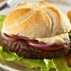 A close-up of a Gourmet Mozzarella Burger on a plate with fresh lettuce, tomato slices, and red onion, all inside a toasted bun. The background includes a casual dining setup with a fork and a glass of juice.