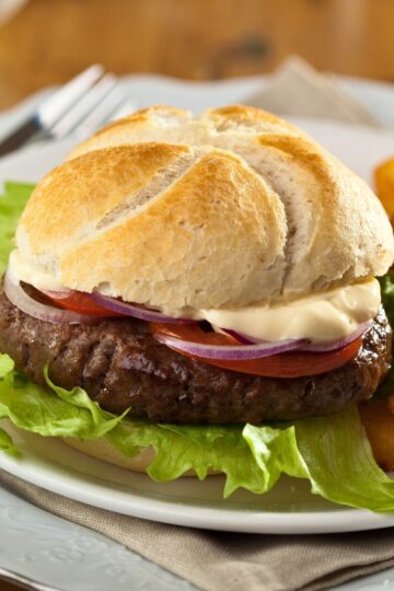 A close-up of a Gourmet Mozzarella Burger on a plate with fresh lettuce, tomato slices, and red onion, all inside a toasted bun. The background includes a casual dining setup with a fork and a glass of juice.