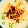Creamy baked potato soup served in a blue bowl, topped with crispy bacon pieces, shredded cheddar cheese, and fresh chopped green onions for a hearty and comforting dish.