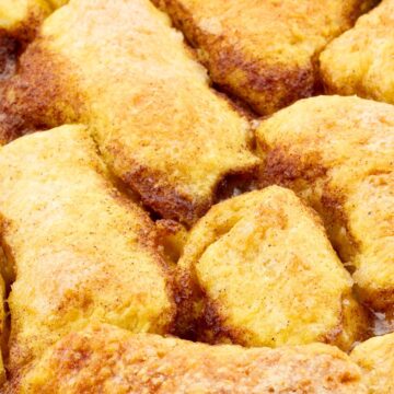 Close-up of golden baked apple dumplings in a casserole dish, coated with cinnamon sugar and bubbling syrup. The flaky pastry is puffed and caramelized, giving a warm, homemade country-style dessert appearance.