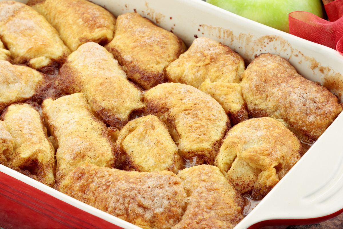 Golden brown classic country apple dumplings baked in a white dish with cinnamon sugar topping and bubbling syrup around the edges, creating a warm and comforting homemade dessert.