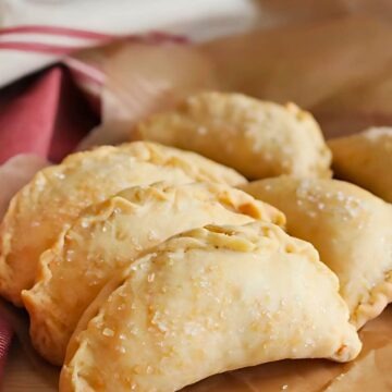 Golden brown cinnamon apple hand pies with flaky crust sprinkled with coarse sugar, arranged on parchment paper. These handheld desserts have a crimped edge and a rustic, homemade look, perfect for fall baking or a sweet treat.