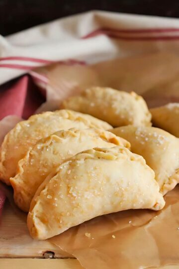 Golden brown cinnamon apple hand pies with flaky crust sprinkled with coarse sugar, arranged on parchment paper. These handheld desserts have a crimped edge and a rustic, homemade look, perfect for fall baking or a sweet treat.
