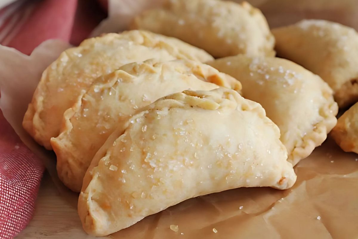 Golden baked cinnamon apple hand pies with crimped edges and a light sprinkle of coarse sugar on top. The flaky pastry shells have a rustic, homemade look, making them a cozy dessert perfect for fall or holiday baking.