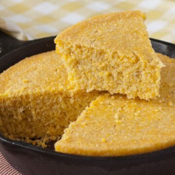 Close-up of sweet skillet cornbread baked in a cast iron pan, cut into wedges with one slice lifted to show the moist, crumbly golden texture. The cornbread has a slightly coarse grain, highlighting the cornmeal base, and sits on a striped cloth with a yellow gingham napkin in the background.