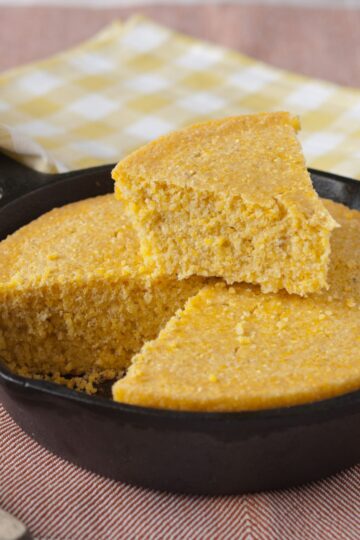 Close-up of sweet skillet cornbread baked in a cast iron pan, cut into wedges with one slice lifted to show the moist, crumbly golden texture. The cornbread has a slightly coarse grain, highlighting the cornmeal base, and sits on a striped cloth with a yellow gingham napkin in the background.
