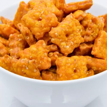 Close-up of a white bowl filled with homemade copycat Cheez-It crackers. The golden orange cheddar squares are baked crisp, topped with coarse salt, and show small center holes, capturing the classic cheesy, crunchy snack look.