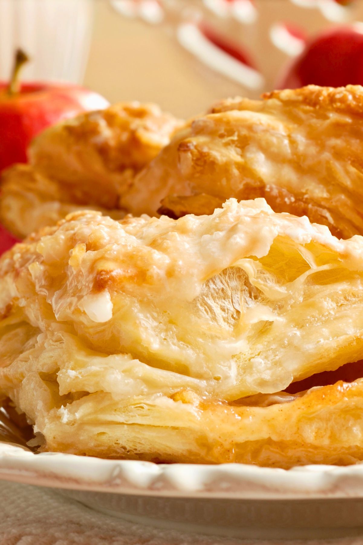 Close-up of a golden, flaky apple turnover topped with a light sugar glaze. The pastry layers are crisp and airy, showing the tender apple filling inside. A white decorative plate holds the turnover, with red apples blurred in the background, giving the scene a warm fall baking feel.