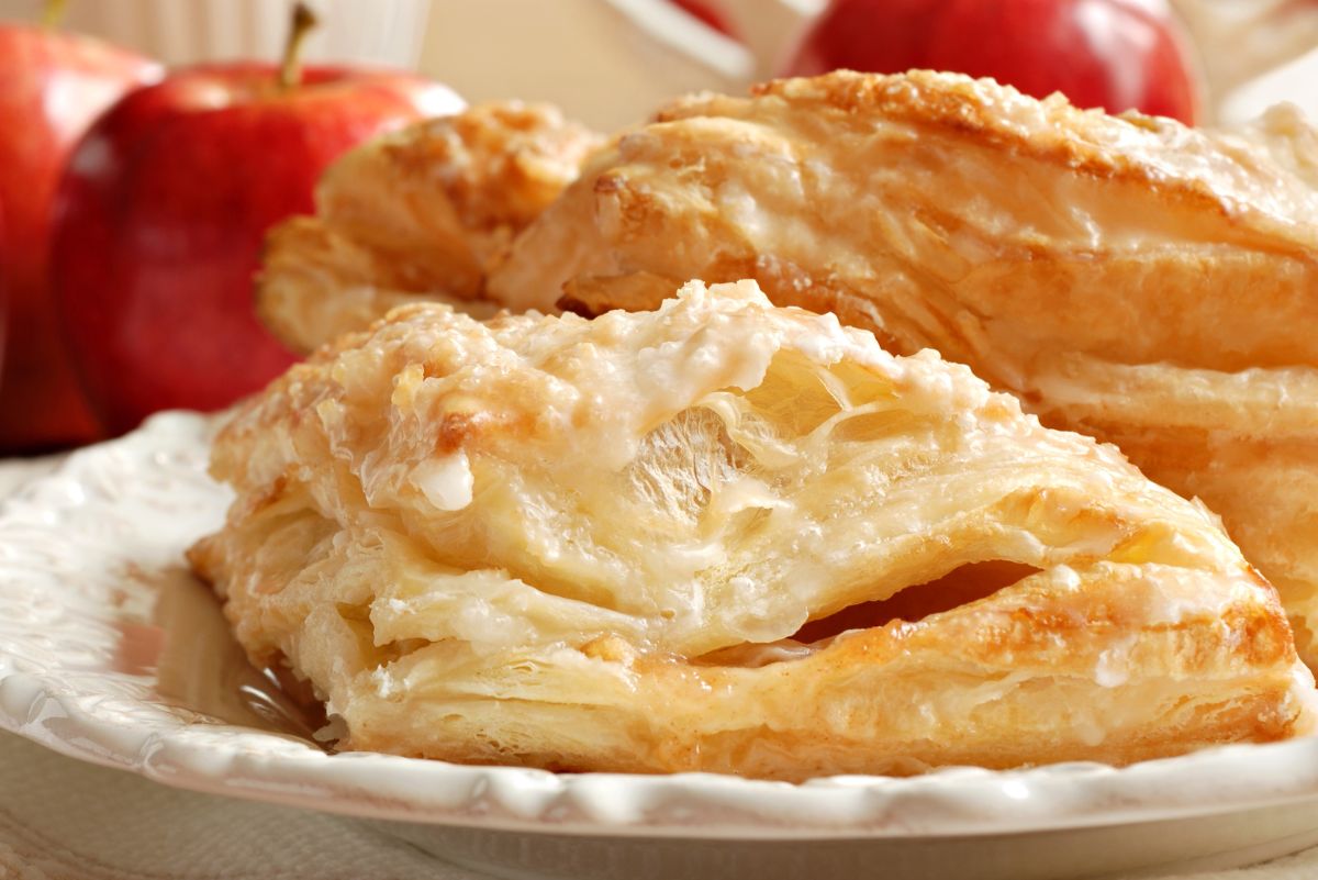 Golden, flaky apple turnovers with a light sugar glaze, served on a decorative white plate. The puff pastry layers are crisp and airy, with baked apple filling inside. Fresh red apples are blurred in the background, emphasizing the fall baking theme.