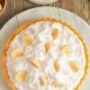 A homemade banana cream pie with a golden flaky crust, topped with fluffy whipped cream and decorated with fresh banana slices. The pie sits on a rustic wooden surface with plates, forks, and bananas nearby, ready to be served.