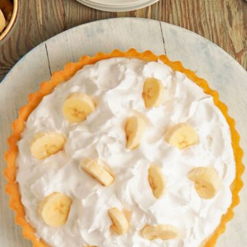 A homemade banana cream pie with a golden flaky crust, topped with fluffy whipped cream and decorated with fresh banana slices. The pie sits on a rustic wooden surface with plates, forks, and bananas nearby, ready to be served.