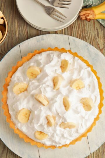 A homemade banana cream pie with a golden flaky crust, topped with fluffy whipped cream and decorated with fresh banana slices. The pie sits on a rustic wooden surface with plates, forks, and bananas nearby, ready to be served.