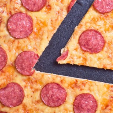 Close-up of a New York style pepperoni pizza with a thin, golden crust and melted cheese. One slice is pulled slightly away, showing the crispy edge and stretchy cheese, highlighting the classic pizzeria look.