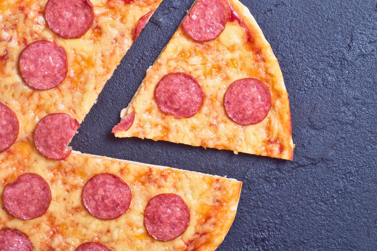 Homemade New York style pepperoni pizza with a thin, golden crust and melted cheese. A single slice is pulled away from the pie, showing the crisp edge and gooey topping against a dark stone surface.