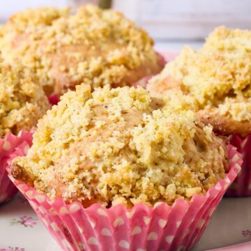 Golden cinnamon apple crumb muffins with buttery streusel topping, baked in pink polka-dot cupcake liners and arranged on a pink plate. Cozy homemade bakery style breakfast or dessert, fall baking recipe, food photography.