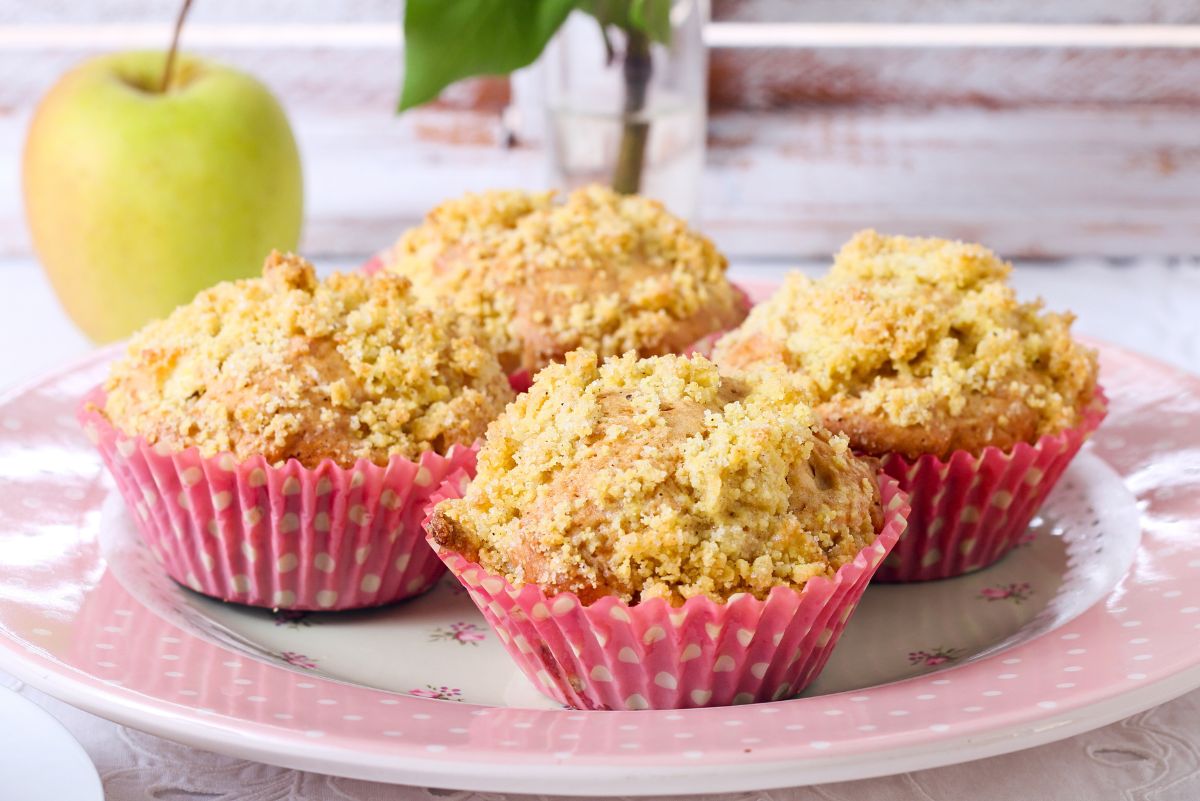 Four cinnamon apple crumb muffins with golden streusel topping sit in pink polka-dot cupcake liners on a pink floral plate. A green apple and small vase blur in the background. Cozy homemade fall baking, breakfast or dessert recipe, food photography.