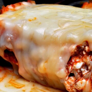 Close-up of Cozy Baked Mexican Lasagna in a casserole dish, a thick blanket of melted cheese over layered tortillas with red salsa-tomato meat sauce and cottage cheese curds visible. Cheesy baked casserole, Mexican lasagna recipe, easy weeknight dinner.