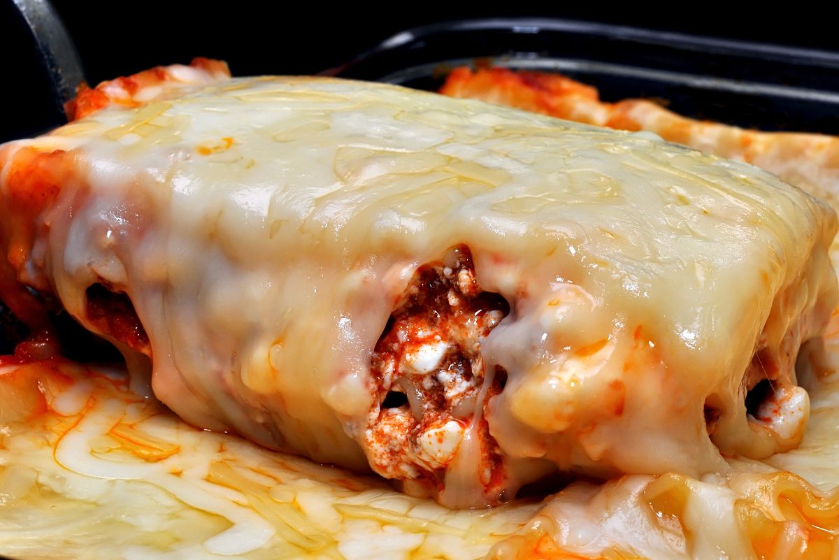Close-up of Cozy Baked Mexican Lasagna: a thick blanket of melted cheese oozes over layered tortillas with red salsa-tomato meat sauce and cottage cheese curds in a casserole dish. Gooey, cheesy comfort food; baked Mexican casserole; easy weeknight dinner recipe.
