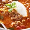 Close-up of quick one-pot lasagna soup in a white bowl: tomato-rich broth with ground beef, curly pasta pieces, and wilted greens, topped with a scoop of ricotta and a fresh basil sprig, spoon at the rim. Cozy Italian dinner recipe.