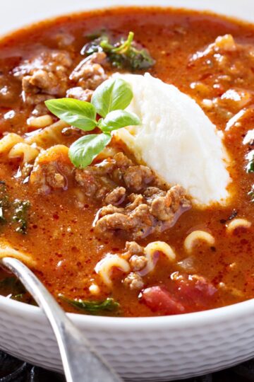 Close-up of quick one-pot lasagna soup in a white bowl: tomato-rich broth with ground beef, curly pasta pieces, and wilted greens, topped with a scoop of ricotta and a fresh basil sprig, spoon at the rim. Cozy Italian dinner recipe.