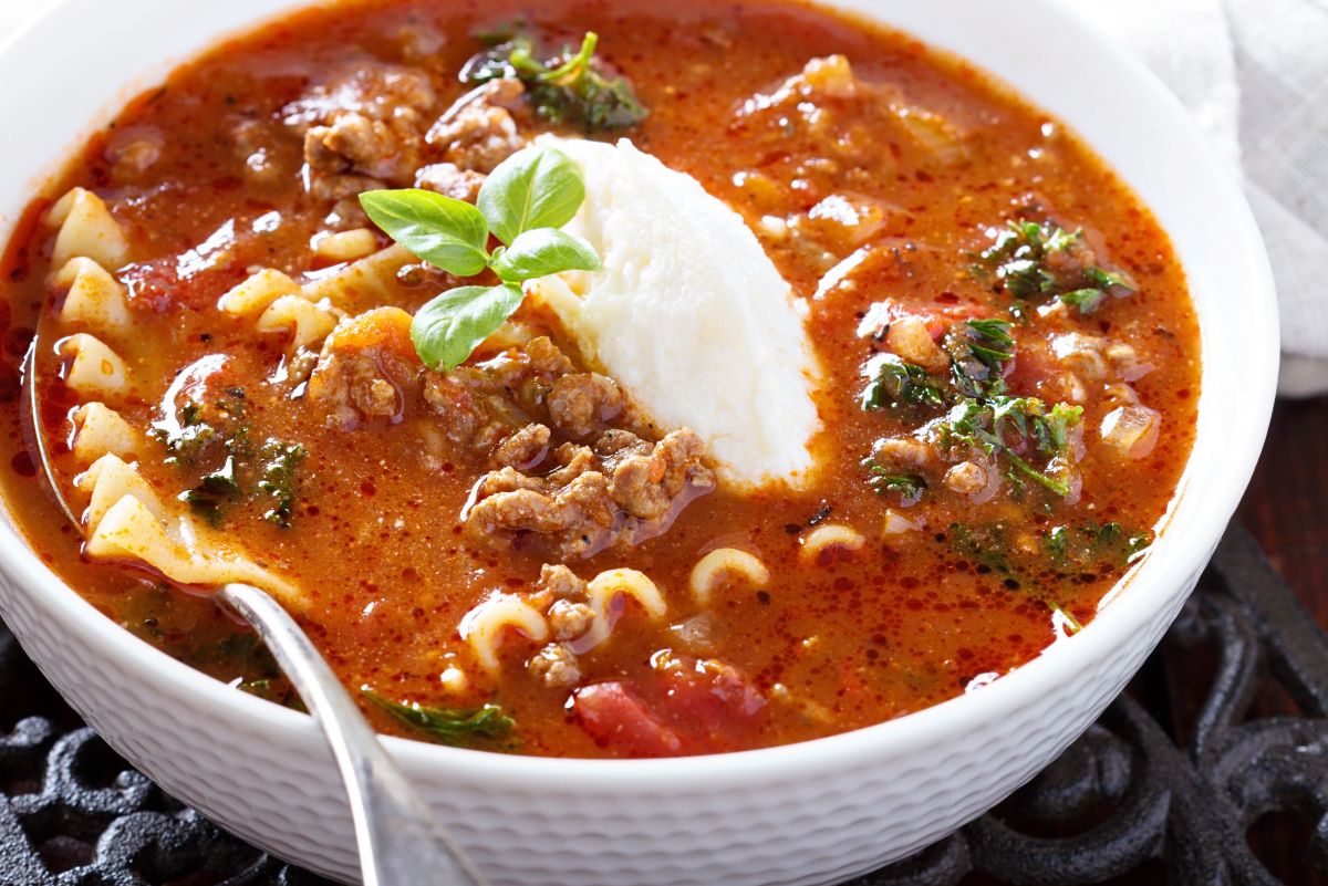 Quick one-pot lasagna soup in a white bowl: rich tomato broth with ground beef, bits of lasagna noodles and curly pasta, wilted greens, and a generous dollop of ricotta topped with fresh basil; spoon at the rim. Cozy Italian weeknight dinner recipe.