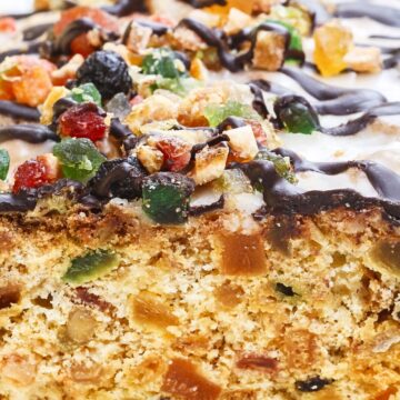Close-up of old-fashioned white Christmas fruitcake, golden crumb packed with candied fruit and nuts, topped with colorful candied pieces and a glossy chocolate drizzle in a white pan. Holiday dessert recipe, fruitcake.