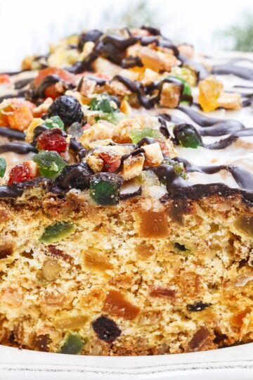 Close-up of old-fashioned white Christmas fruitcake, golden crumb packed with candied fruit and nuts, topped with colorful candied pieces and a glossy chocolate drizzle in a white pan. Holiday dessert recipe, fruitcake.