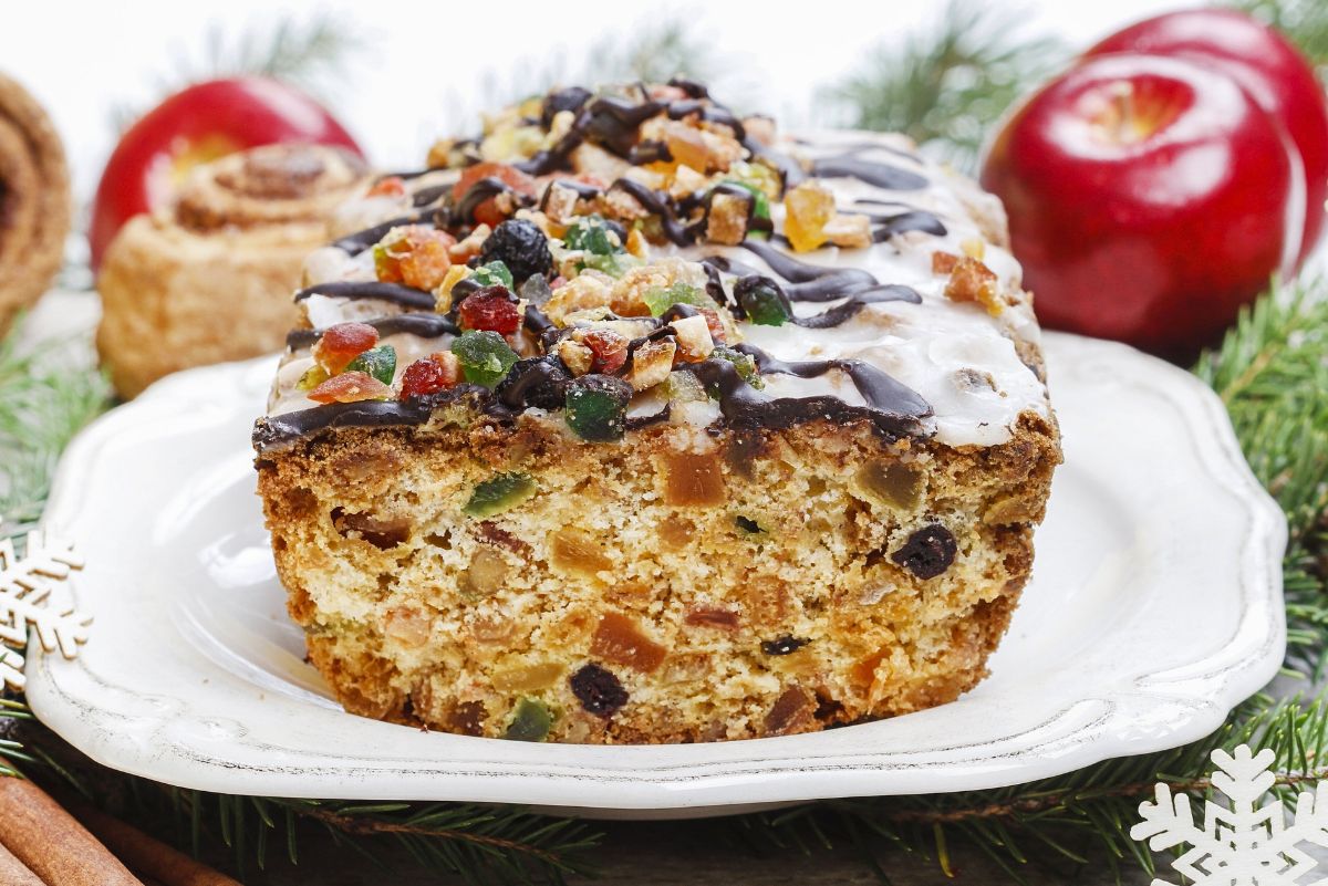 Old-fashioned white Christmas fruitcake loaf on a white platter, golden crumb packed with candied fruit and nuts, topped with icing and chocolate drizzle. Holiday dessert recipe with evergreen sprigs, apples, and cinnamon sticks.