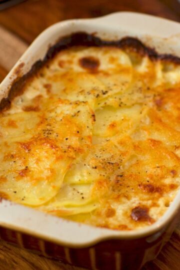 Golden baked potato lasagna in a white casserole dish with creamy Alfredo sauce and melted cheese on top, lightly browned and sprinkled with black pepper. The edges of the dish show caramelized sauce from baking, giving it a cozy, homemade look.