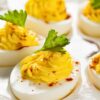 Close-up of spicy deviled eggs with smooth, piped yellow yolk filling made with hot pepper sauce, lightly dusted with paprika and topped with fresh parsley leaves. The deviled eggs are arranged on a white serving platter with a clean, bright background, highlighting a classic appetizer perfect for parties and holidays.
