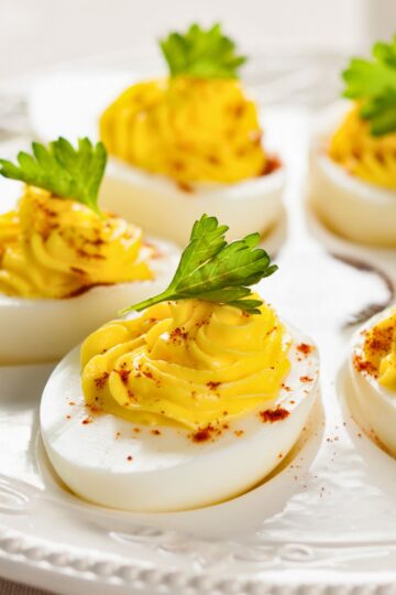 Close-up of spicy deviled eggs with smooth, piped yellow yolk filling made with hot pepper sauce, lightly dusted with paprika and topped with fresh parsley leaves. The deviled eggs are arranged on a white serving platter with a clean, bright background, highlighting a classic appetizer perfect for parties and holidays.