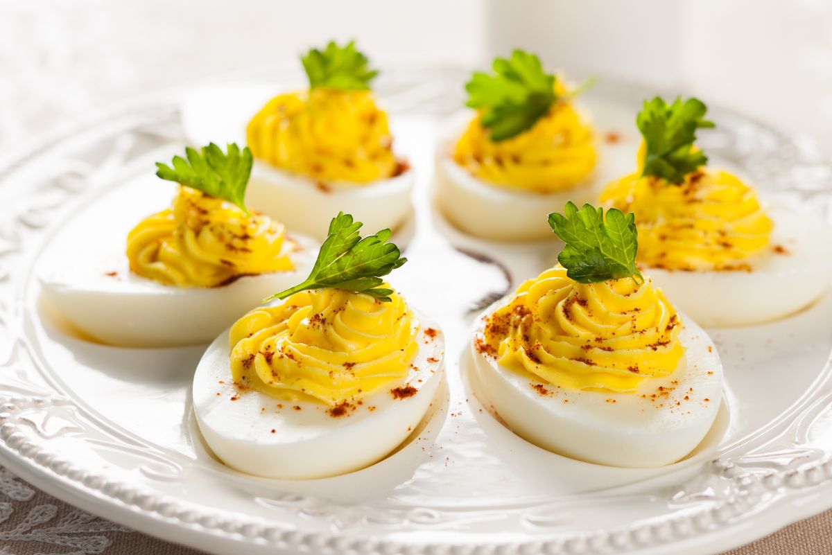Spicy deviled eggs arranged on a white decorative platter, each egg half filled with creamy piped yolk made with hot pepper sauce, sprinkled with paprika and topped with fresh parsley. Bright yellow filling contrasts with the white eggs for a classic deviled egg appetizer presentation.