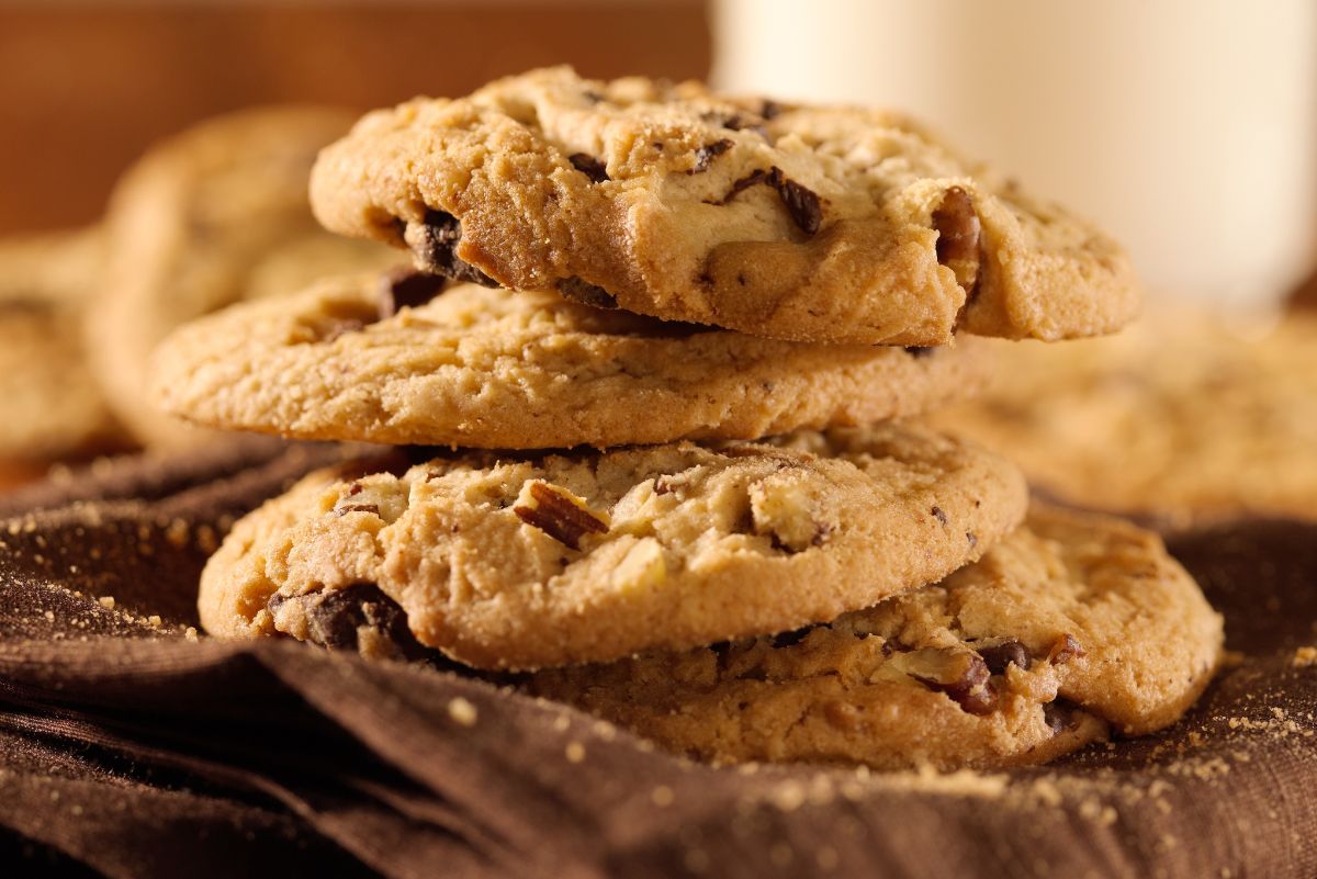 A cozy stack of golden chocolate chip hazelnut cookies resting on a brown cloth, with crumbs scattered around. The cookies have a crisp edge, soft centers, visible chocolate chunks, and chopped hazelnuts, giving a warm homemade bakery feel.