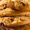 Warm, golden chocolate chip hazelnut cookies stacked together, showing a crumbly texture, toasted hazelnut pieces, and melted chocolate chips. Soft browns and cozy lighting give a fresh-baked kitchen vibe.