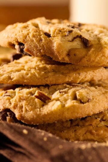 Warm, golden chocolate chip hazelnut cookies stacked together, showing a crumbly texture, toasted hazelnut pieces, and melted chocolate chips. Soft browns and cozy lighting give a fresh-baked kitchen vibe.