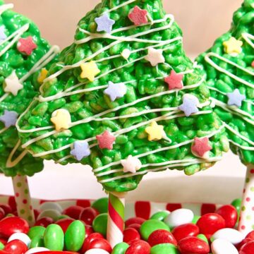 Bright green Christmas tree Rice Krispies treat on a striped stick, drizzled with white icing and topped with pastel star sprinkles. It stands in a festive red and green dish filled with red, green, and white candies for a fun holiday dessert display.