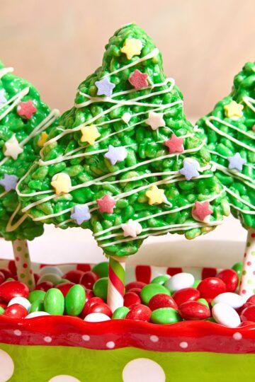 Bright green Christmas tree Rice Krispies treat on a striped stick, drizzled with white icing and topped with pastel star sprinkles. It stands in a festive red and green dish filled with red, green, and white candies for a fun holiday dessert display.