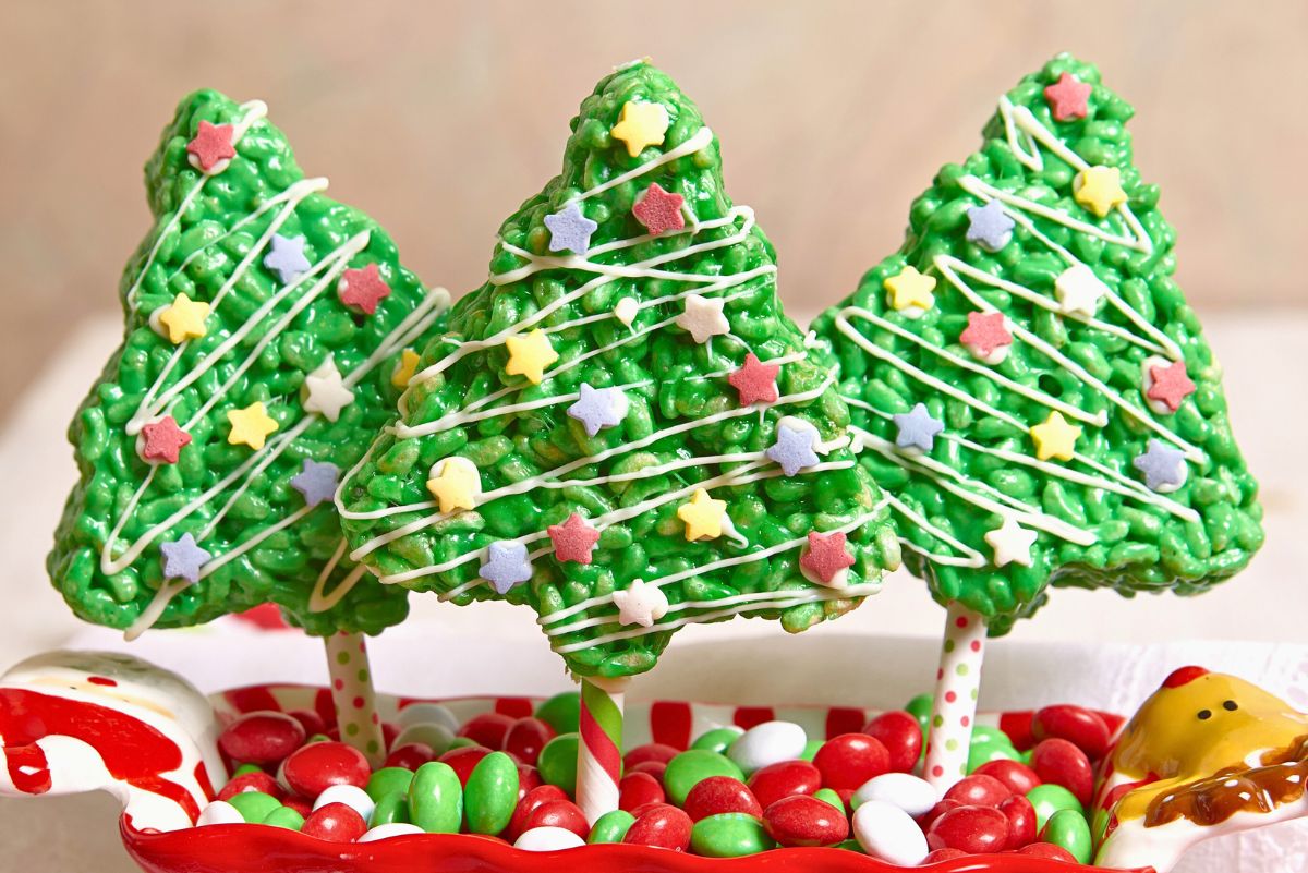 Three green Christmas tree Rice Krispies treats on festive striped sticks, drizzled with white icing and decorated with colorful star sprinkles. They stand in a red and green dish filled with holiday candies for a bright, cheerful Christmas dessert display.