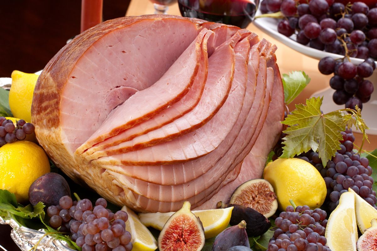 Spiral-sliced honey baked ham displayed on a silver platter surrounded by fresh grapes, figs, and lemon wedges. The glossy pink ham slices fan out in the center, creating a festive holiday ham presentation perfect for a baked ham recipe or Christmas dinner spread.