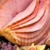 Spiral-sliced honey baked ham displayed on a platter with glossy pink slices, surrounded by fresh figs, lemon wedges, and a small cluster of purple grapes. Warm, festive presentation ideal for holiday ham or baked ham recipe