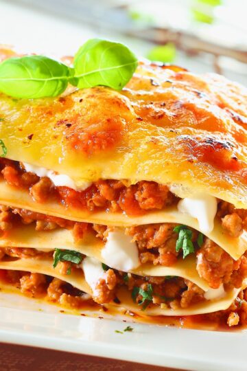 Cheesy beef lasagna with golden bubbly top, stacked pasta layers, rich meat sauce, creamy ricotta, and fresh basil on top.