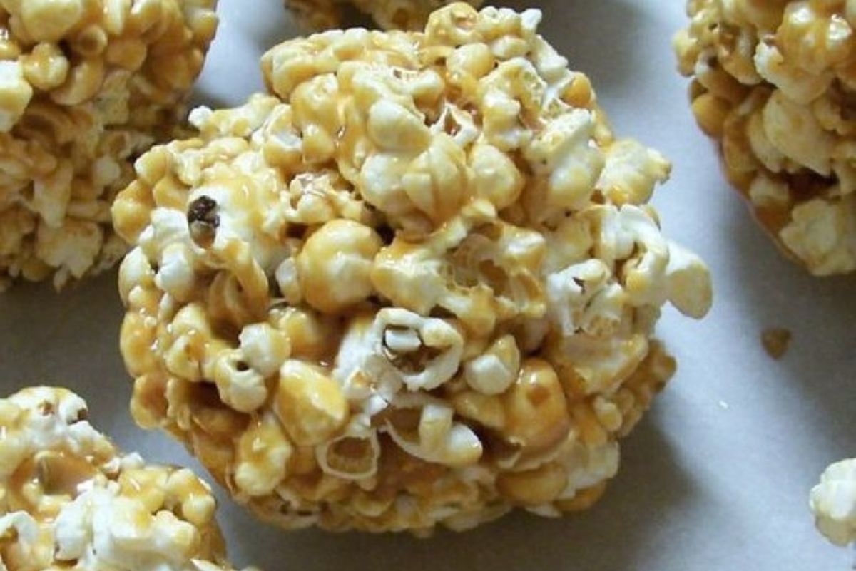 Close-up of a homemade popcorn ball coated in golden caramel on a white surface. Fluffy popped corn bound together with glossy caramel, showing a crunchy texture and uneven, rustic shape. Sweet popcorn dessert snack photographed from above.