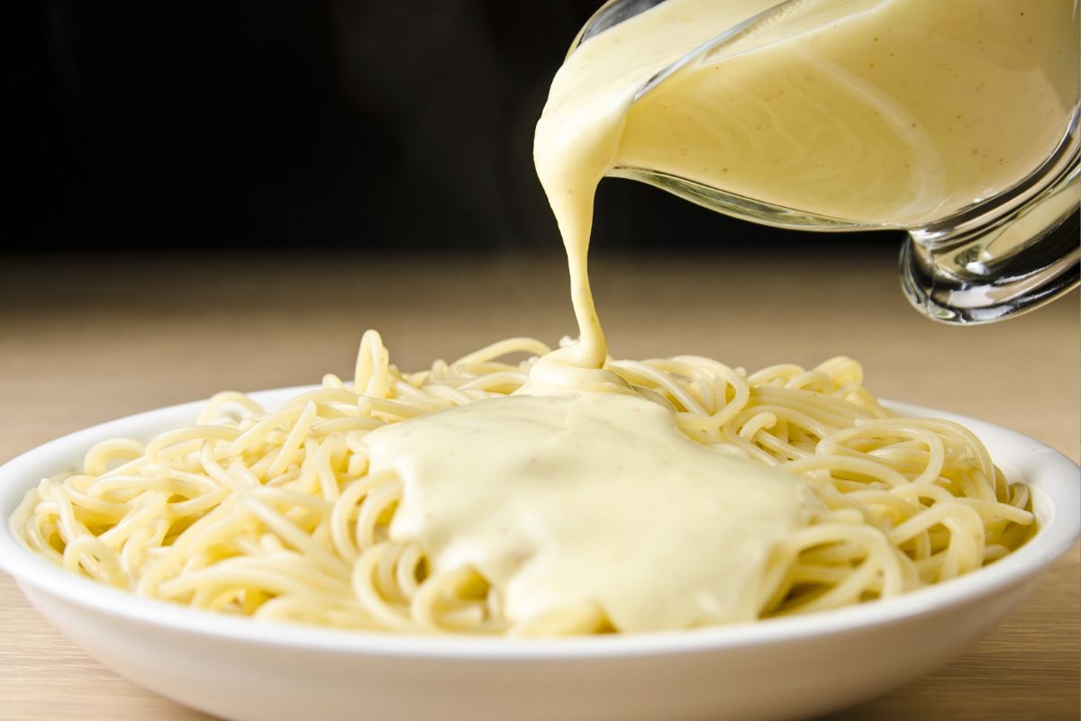 Creamy four cheese pasta sauce pouring from a glass measuring cup over a plate of spaghetti in a white bowl. Thick, smooth white cheese sauce coats the noodles. Quick 15 minute homemade cheese pasta sauce for easy dinner.