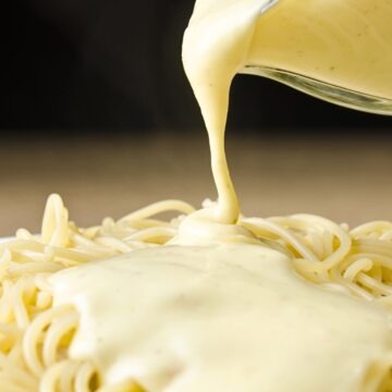 Close-up of creamy four cheese pasta sauce pouring from a glass measuring cup onto spaghetti. Thick, smooth white cheese sauce coats the noodles in a white bowl. Quick 15 minute homemade cheese pasta sauce.