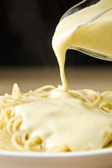 Close-up of creamy four cheese pasta sauce pouring from a glass measuring cup onto spaghetti. Thick, smooth white cheese sauce coats the noodles in a white bowl. Quick 15 minute homemade cheese pasta sauce.